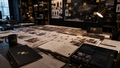 A dark, atmospheric tabletop scene in a studio or office, with sketches, notebooks, a tablet, and a mug arranged on a desk under a lit lamp. No person is visible, but the real environment is present, so this fits Lifestyle rather than Product/Packaging.