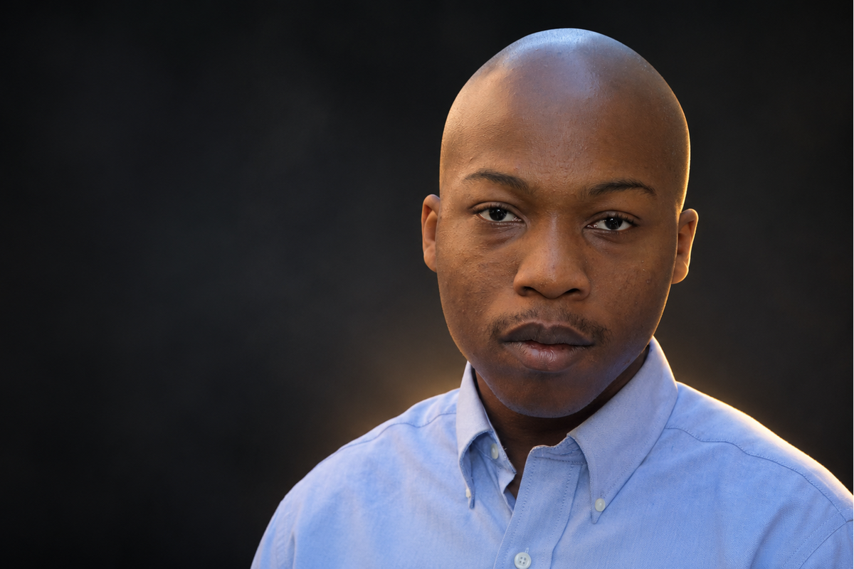 A close-up portrait of a bald man wearing a light blue button-down shirt, set against a dark, moody background with a warm spotlight on his face.