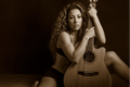 A woman with curly hair sits on a dark studio floor holding and cradling an acoustic guitar. The photo is rendered in a warm sepia tone, with the model wearing a black bra and shorts.
