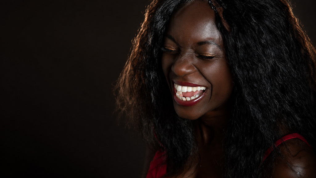 A close-up portrait of a smiling woman with long, wavy black hair, wearing a red top against a dark background.